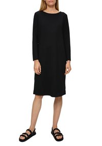 Schwarzes, langärmeliges Kleid aus strukturiertem Stoff, mit Rundhalsausschnitt und lockerer Passform, kombiniert mit schwarzen Sandalen.