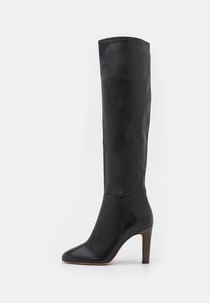 Botte en cuir noire montant jusqu'aux genoux avec bout pointu et talon haut empilé sur fond blanc.