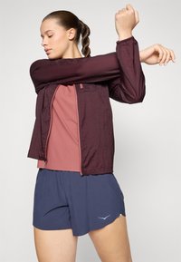 Nainen marsalkanpunaisessa takissa, vaaleanpunaisessa topissa ja laivastonsinisissä shortseissa venyttää kättään rinnan yli yksiväristä taustaa vasten.