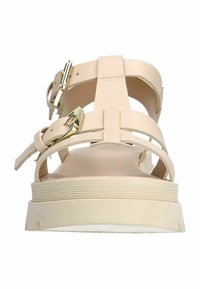 Beige sandaal met kruisbandontwerp, verstelbare goudkleurige gespen en een dikke, gestructureerde zool met horizontale ribbels.