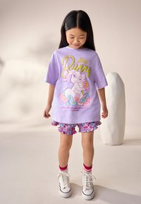 Lila T-Shirt mit "Miss Bunny"-Grafik, kombiniert mit geblümten Shorts mit Rüschen. Strukturierte weiße High-Top-Sneaker runden den Look ab.