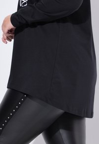 Schwarzes Langarmshirt mit rundem Ausschnitt, kombiniert mit schwarzen Leggings, die entlang der Seite eine dekorative vertikale Reihe von silbernen Akzenten aufweisen.