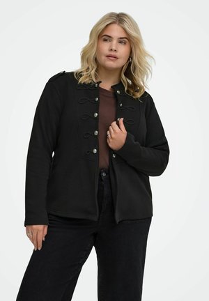 Blond kvinde iført en sort jakke med dekorative knapper over en brun skjorte og sorte bukser, stående mod en ensfarvet hvid baggrund.