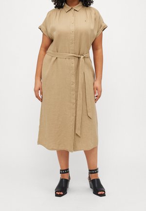 Kvinde iført en beige midikjole med knapper, taljebånd og korte ærmer, kombineret med sorte åbne tå-sandaler med ankelrem.