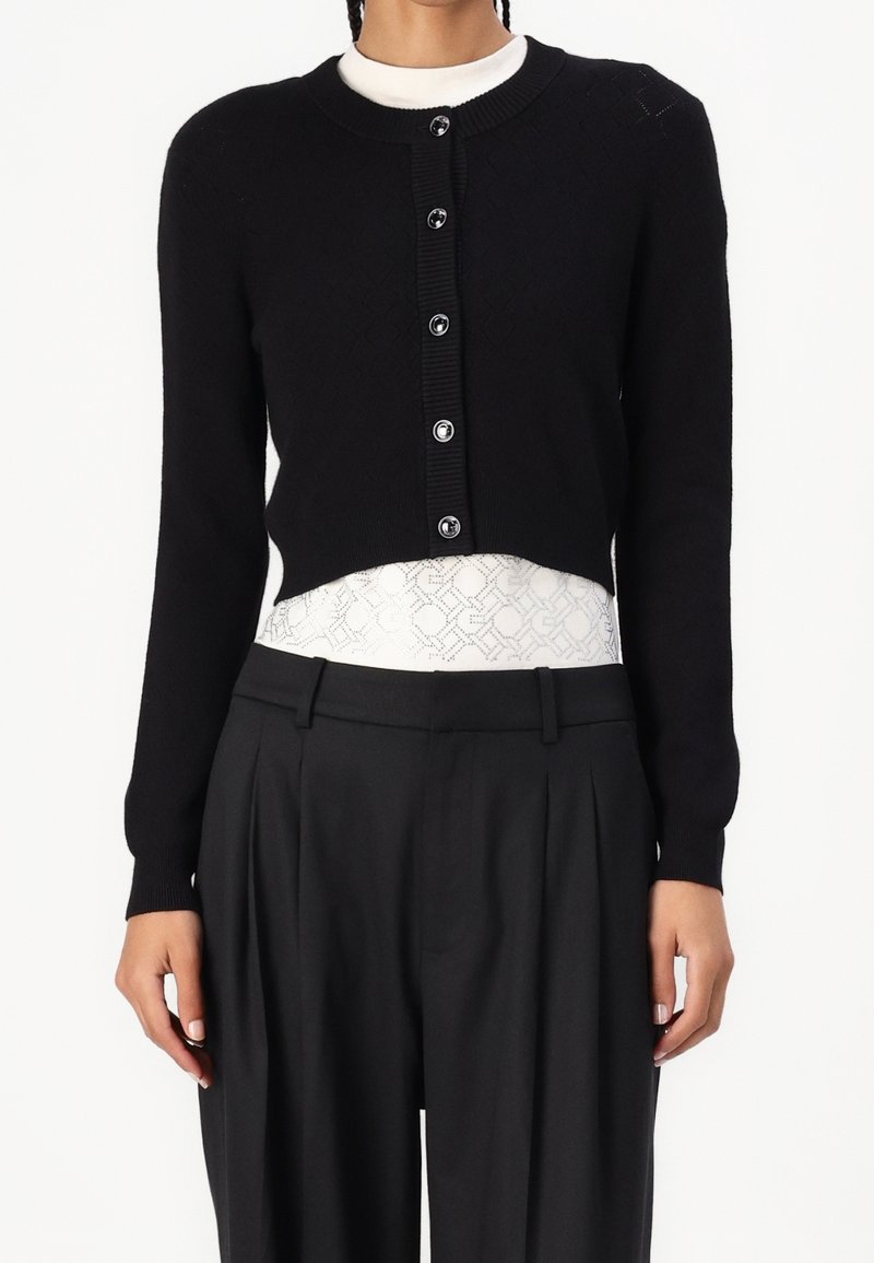 Femme portant un cardigan court noir boutonné sur un top blanc à motifs, associé à un pantalon noir plissé taille haute.