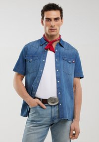 Camicia di jeans a maniche corte con bottoni a pressione, due tasche sul petto e yoke appuntito. Indossata sopra una maglietta bianca, abbinata a jeans azzurri chiari. Accento di bandana rossa.
