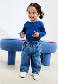 Plava košulja dugih rukava u kombinaciji s traper hlačama s elastičnim strukom i uzorkom medvjedića. Dijete nosi bijele tenisice, stojeći u neutralnom prostoru.