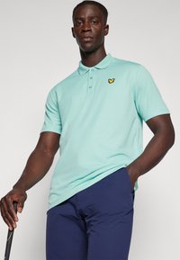 Lys grønn polo skjorte med krage og tre knapper, prydet med en liten gul sommerfugl-logo på venstre bryst. Bæres med marineblå bukser.
