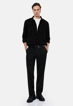 Mand stående med hænderne i lommerne iført sort skjorte med knapper, sorte bukser, sort bælte, sorte sko og hvid undertrøje på hvid baggrund.