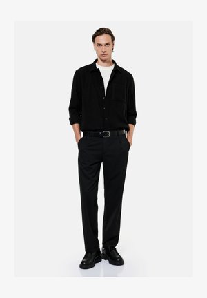 Mand stående med hænderne i lommerne iført sort skjorte med knapper, sorte bukser, sort bælte, sorte sko og hvid undertrøje på hvid baggrund.