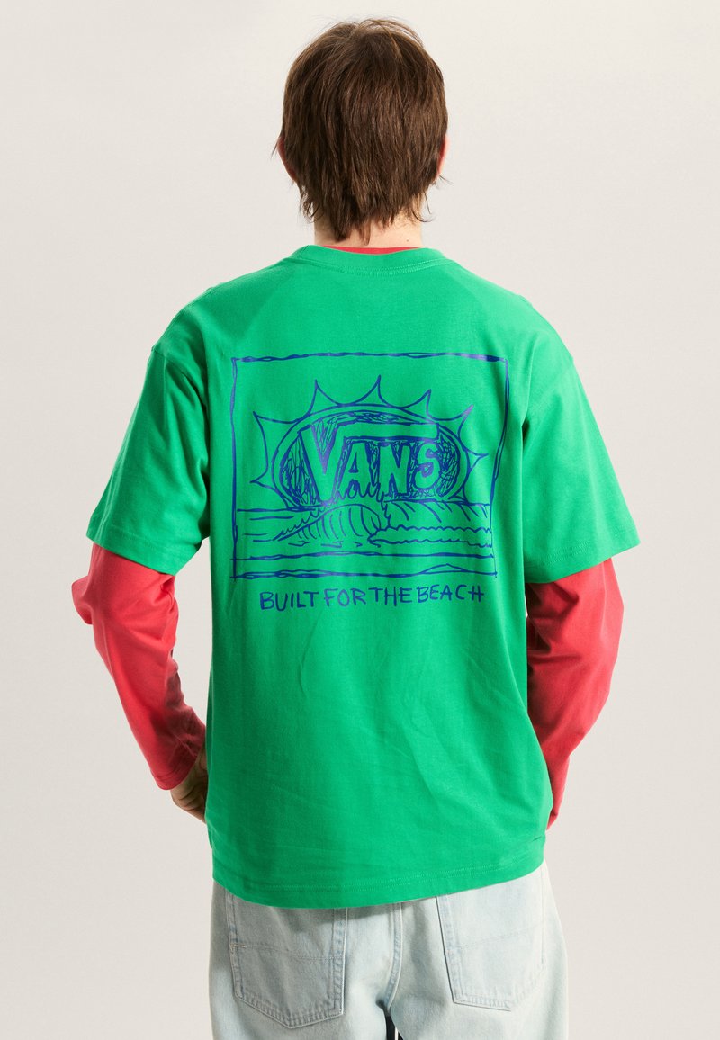 T-shirt en coton vert avec un motif graphique bleu affichant les mots « VANS » et « BUILT FOR THE BEACH », porté par-dessus un t-shirt rouge à manches longues.