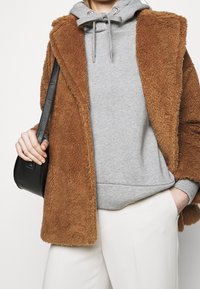 Persoon in een bruine textuurjas over een grijze hoodie, witte broek, met een zwarte handtas in de hand en de andere hand in de zak.