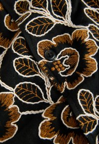 Musta kangas, jossa on ruskeita ja valkoisia kirjottuja kukkakuoseja, ja keskellä on ommeltu musta nelireikäinen nappi.
