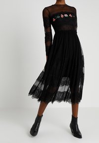 Robe en dentelle noire avec un haut transparent à manches longues orné de broderies florales, un corsage ajusté et une jupe fluide à couches. Bottines incluses.
