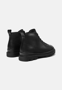 Botas de tobillo de cuero negro con punta redondeada, cordones en la parte delantera, cremallera trasera y suela gruesa texturizada con pequeñas protuberancias.