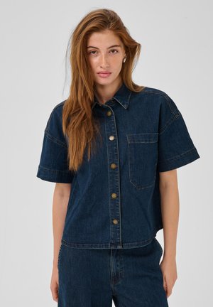 Jeune femme aux longs cheveux roux portant une chemise en jean bleu foncé à manches courtes et un pantalon en jean assorti sur un fond clair uni.