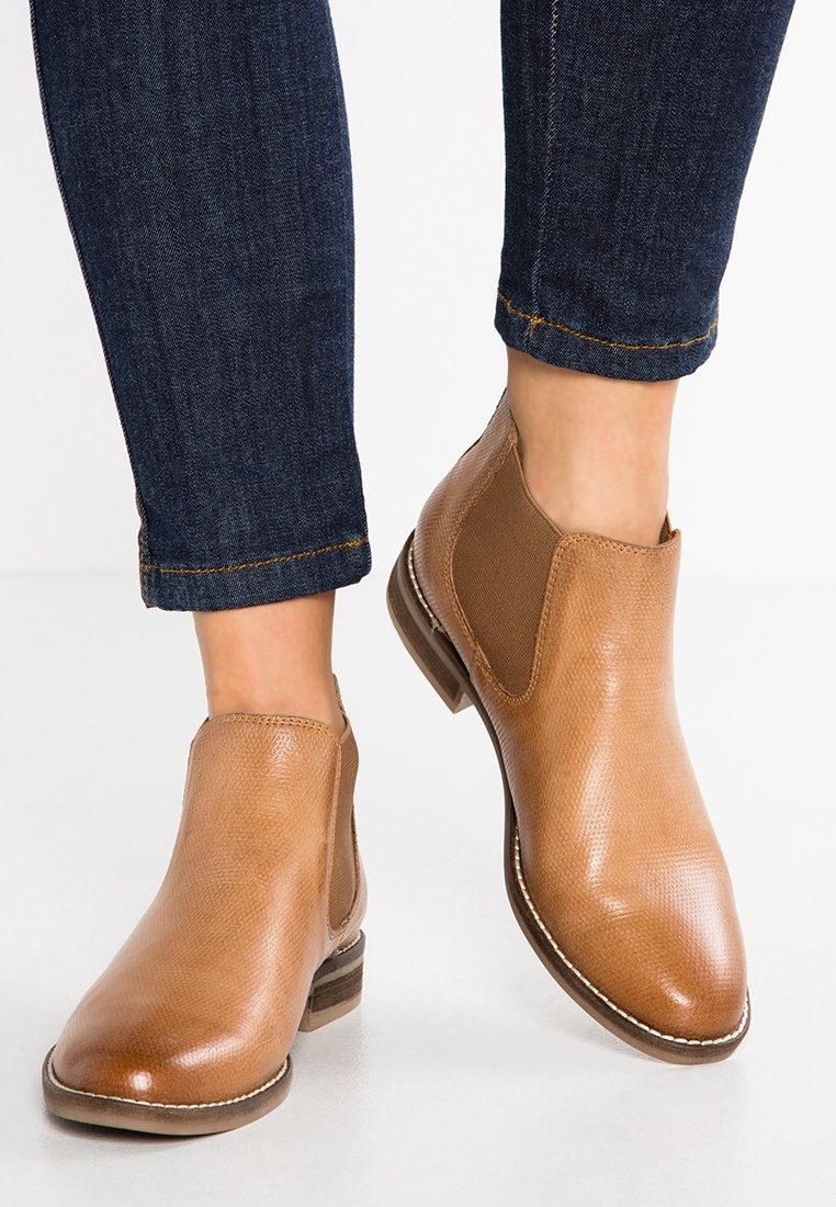 Bottes chevilles en cuir fauve avec talons empilés bas portées avec un jean bleu foncé retroussé sur fond blanc.