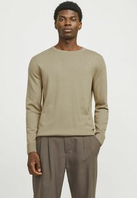 Beige gestrickter Pullover mit Rundhalsausschnitt und langen Ärmeln, kombiniert mit lockeren braunen Hosen. Mit gerippten Bündchen und Saum, glatte Textur.