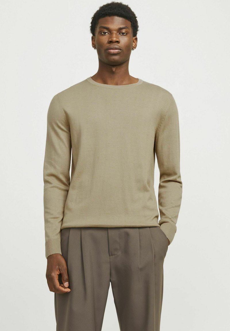 Beige gestrickter Pullover mit Rundhalsausschnitt und langen Ärmeln, kombiniert mit lockeren braunen Hosen. Mit gerippten Bündchen und Saum, glatte Textur.