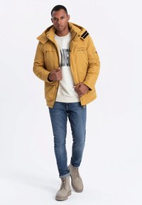 Gul puffjacka med huva, med svarta detaljer, tillsammans med en ljus tröja, blå jeans och beige snörkängor.