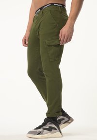 Groene cargobroek met een aansluitend ontwerp, rekbaar materiaal, zijzakken en een zwarte tailleband met de tekst "STHUGE". Gecombineerd met grijs en zwarte sneakers.