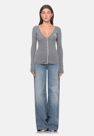 Donna che indossa un cardigan grigio aderente con bottoni, con scollo a V profondo e jeans blu a gamba larga, in piedi davanti a uno sfondo semplice.