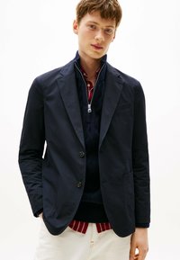 Blazer blu navy realizzato in tessuto strutturato, con due bottoni, tasche con pattina e vestibilità sagomata. Indossato sopra un maglione con cerniera e una camicia con colletto.