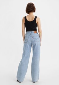Femme aux cheveux courts bruns portant un haut noir sans manches et un jean large taille haute bleu clair, debout de dos.