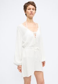 Γυναίκα με κοντά μαλλιά φοράει ένα λευκό, μακρυμάνικο φόρεμα με V λαιμόκοψη, ζώνη στη μέση και φαίνεται λουράκι χαλτερ κάτω από τη λαιμόκοψη.