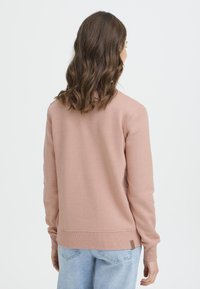 Jasnoróżowa bluza z długimi rękawami, przeszywanym dołem i małą materiałową metką na dolnej lewej stronie. Miękka, gładka faktura, luźny krój.