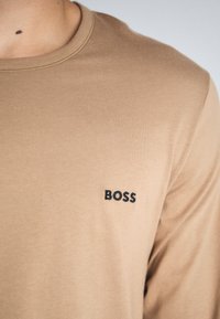 Šviesiai rudas ilgo rankovėmis marškinėliai su apvalia iškirpte ir mažuoju mustruotu juodu "BOSS" logotipu kairėje krūtinės pusėje. Lygus audinio tekstūra.