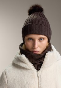 Hnedá pletená čiapka s pom-pomom na vrchu, skombinovaná s rovnakou hnedou rebrovanou šálou. Model má na sebe nadýchanú krémovú bundu.