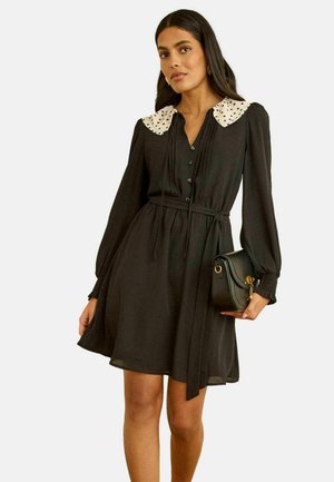 Kvinde iført en sort kjole med lange ærmer og en krave med hvide polkaprikker, holder en sort håndtaske og står foran en ensfarvet baggrund.