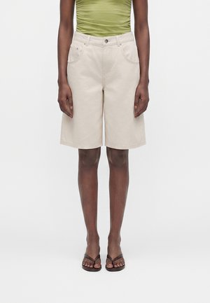Person iført beige knælange shorts, olivengrøn ærmeløs top og sorte flip-flops stående mod hvid baggrund.