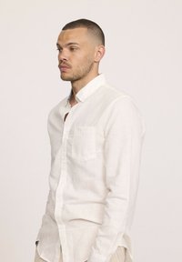 Chemise en lin blanc à boutons avec une poche, présentant une texture subtile et un col décontracté, associée à un pantalon de teintes neutres.