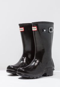 Bottes en caoutchouc noires, imperméables, avec une finition brillante, un accent en boucle et des étiquettes rouges affichant le nom de la marque sur les côtés extérieurs.