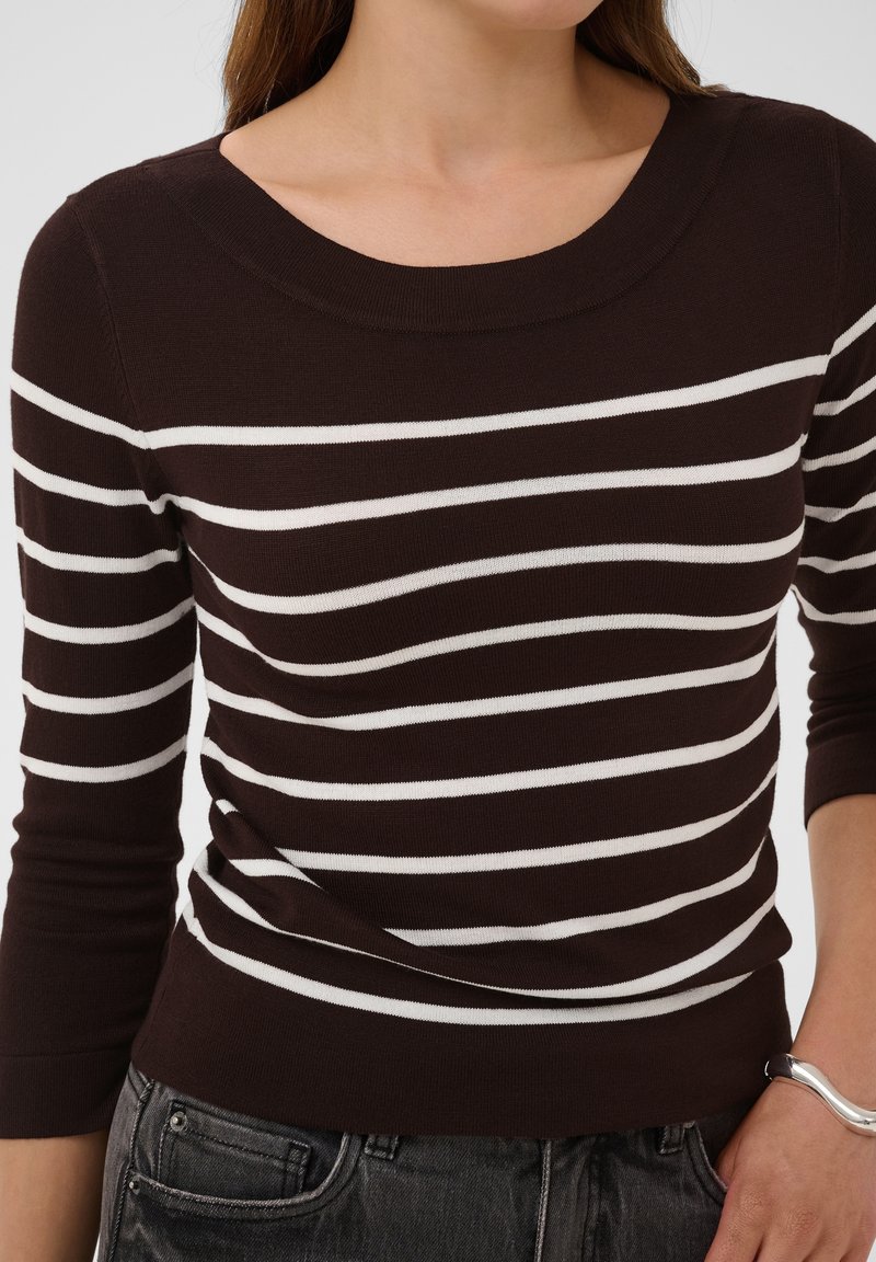 Femme portant un pull marron foncé avec des rayures horizontales blanches et des manches trois-quarts, associé à un jean gris foncé et un bracelet argenté.
