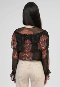 Blouse noire transparente avec des couches froncées, ornée d'un motif floral marron et crème. Manches longues avec des poignets froncés.
