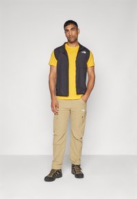 Schwarze ärmellose Weste über einem gelben T-Shirt, kombiniert mit beige Cargo-Hosen und schwarzen Wanderschuhen mit orangen Akzenten.