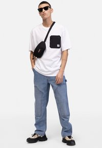 White t-shirt with a black pocket, blue jeans with curved seams, black crossbody bag, and black sunglasses. Footwear features beige and black accents.