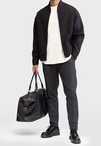 Homme en veste noire, chemise blanche, pantalon gris, chaussures noires tenant un grand sac de voyage en cuir noir contre un fond clair.