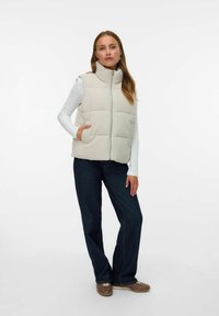 Beige gewatteerde vest met hoge kraag, zijzakken en een ritsluiting, gedragen over een witte langekleding top en gecombineerd met donkere jeans.