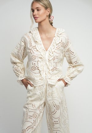 Blonde vrouw die een crèmekleurige ajour kanten blouse met ruches draagt en bijpassende wijde broek, handen in de zakken, kijkt iets naar links.