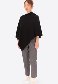 Poncho nero in tessuto morbido, con un design drappeggiato e un orlo asimmetrico, abbinato a pantaloni grigi chiari a sigaretta e sneakers bianche.