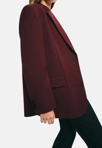 Kvinna klädd i en mörkröd skräddarsydd blazer med svarta knappar och en ficklock, tillsammans med svarta kostymbyxor med ränder, på en vit bakgrund.