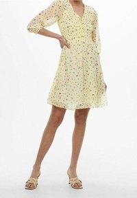 Vaaleankeltainen mekko, jossa on kukkakuosisia, kootut rinnan osat ja läpikuultavat hihat. Mekko laskeutuu löysään hameeseen. Yhdistettynä beigeihin korokepantoihin.