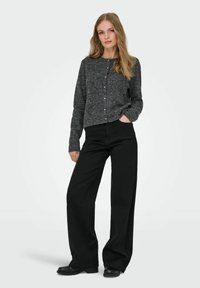 En ung kvinna står med en mörkgrå, knäppt tröja, vida svarta byxor och svarta skor, med en hand i fickan.