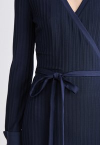 Navy blue wrap dress with long sleeves and ribbed texture. Features a tied waist and a deep V-neckline, offering structured design.