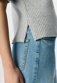 Maglione grigio a coste con fessure laterali, abbinato a jeans in denim azzurro chiaro, che mostrano una texture liscia e minimi dettagli di cucitura.