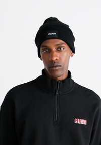 Μαύρο μπιμπελό με διπλωμένο γείσο και λευκή ετικέτα λογότυπου, σε συνδυασμό με μαύρο φούτερ με φερμουάρ στο λαιμό που διαθέτει κόκκινο κεντημένο λογότυπο στο στήθος.